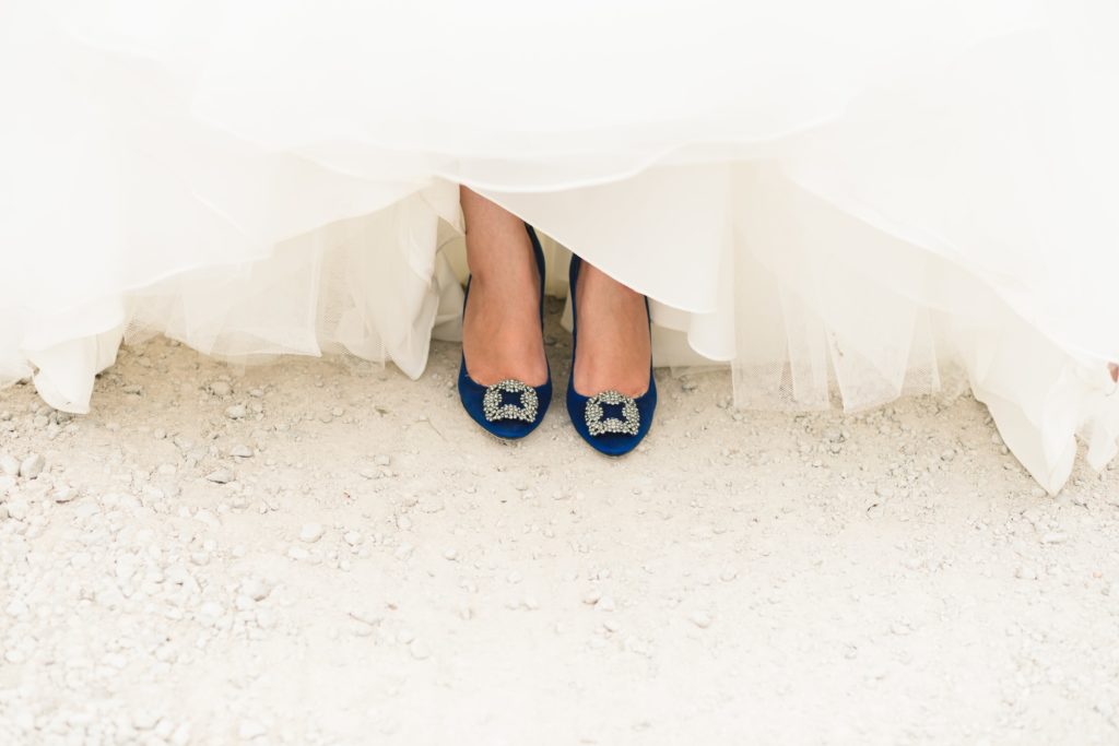 A bride stands with their dress slightly pulled up to reveal their blue, embroidered shoes.
